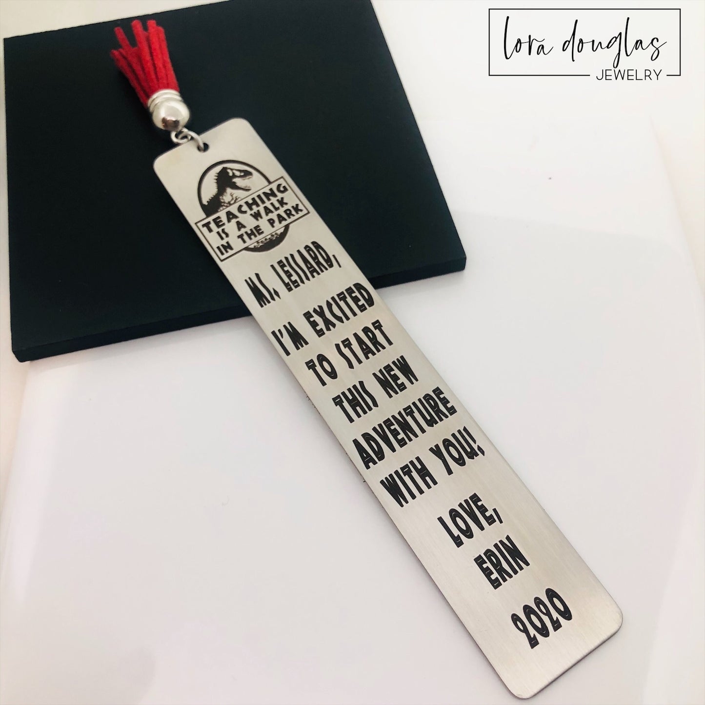 A stainless steel bookmark with an engraved message for a teacher, accompanied by a tassel and packaged for gift giving