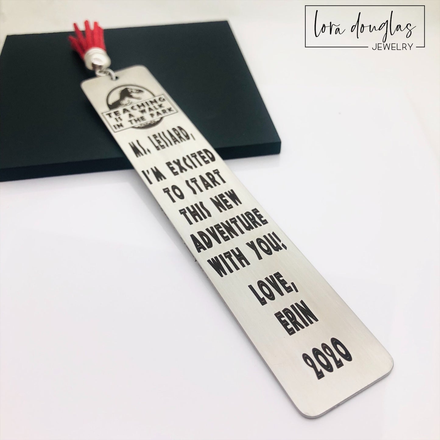 A stainless steel bookmark with an engraved message for a teacher, accompanied by a tassel and packaged for gift giving