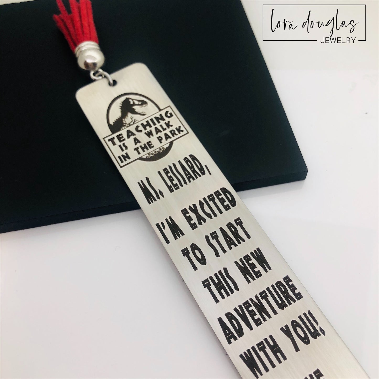 A stainless steel bookmark with an engraved message for a teacher, accompanied by a tassel and packaged for gift giving