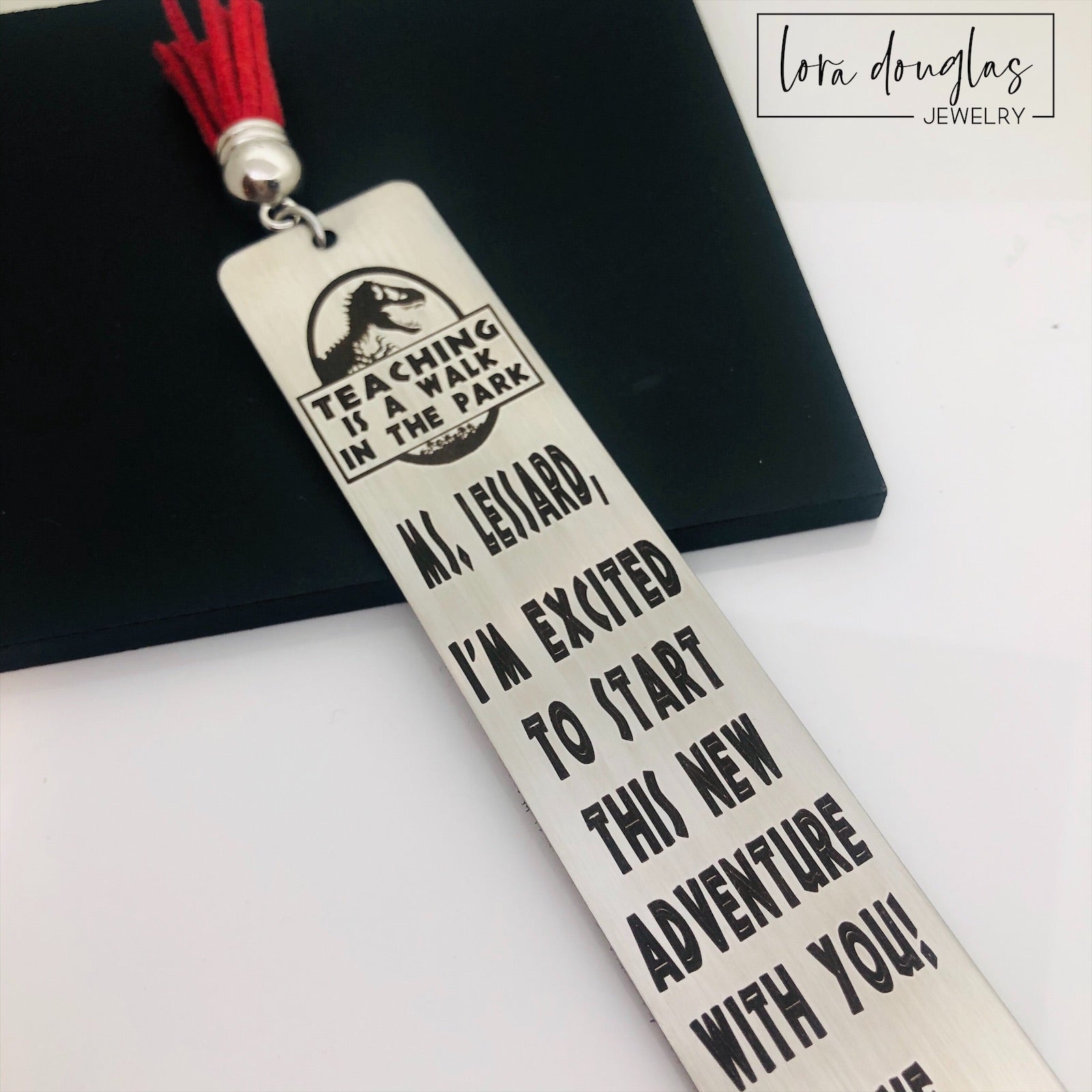 A stainless steel bookmark with an engraved message for a teacher, accompanied by a tassel and packaged for gift giving