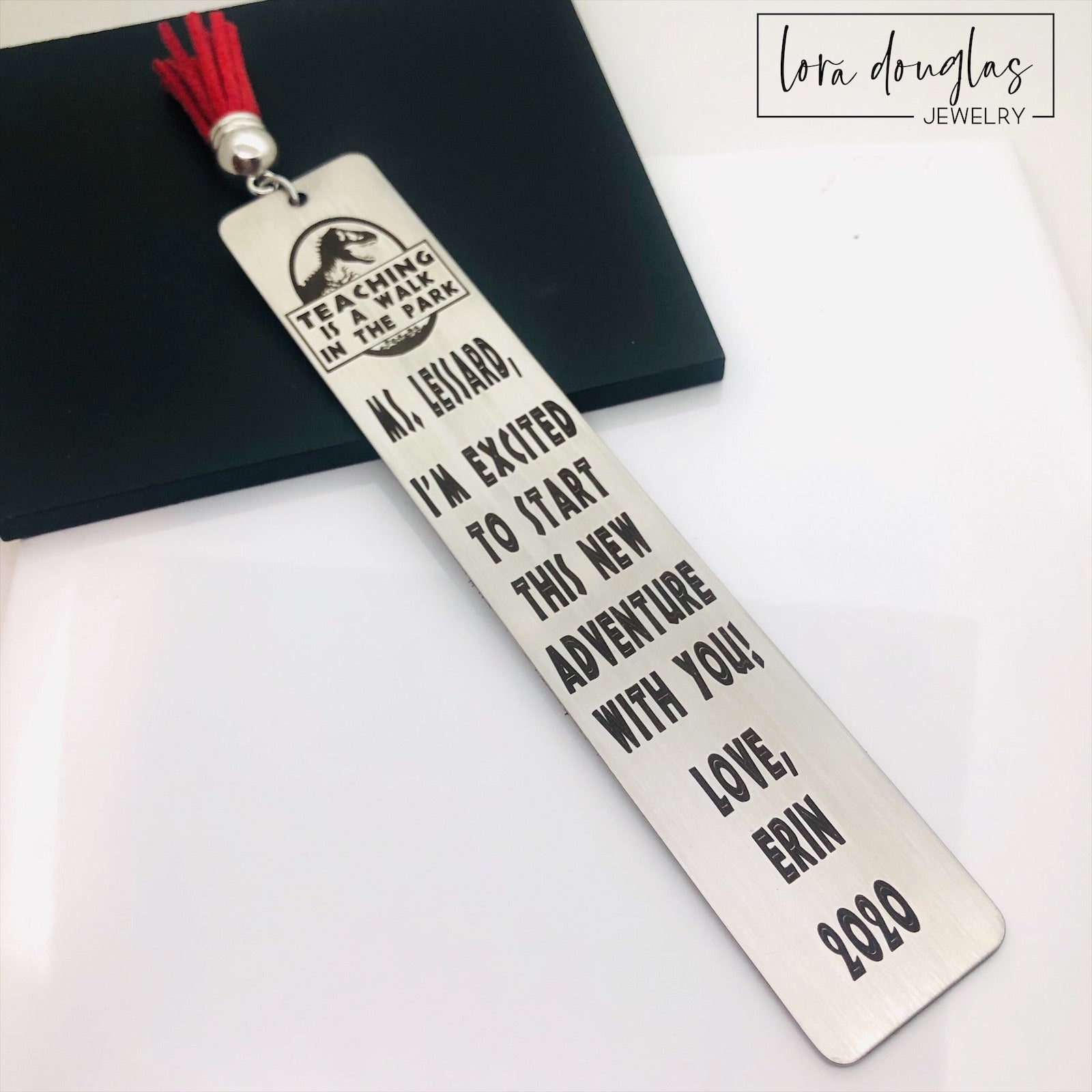 A stainless steel bookmark with an engraved message for a teacher, accompanied by a tassel and packaged for gift giving