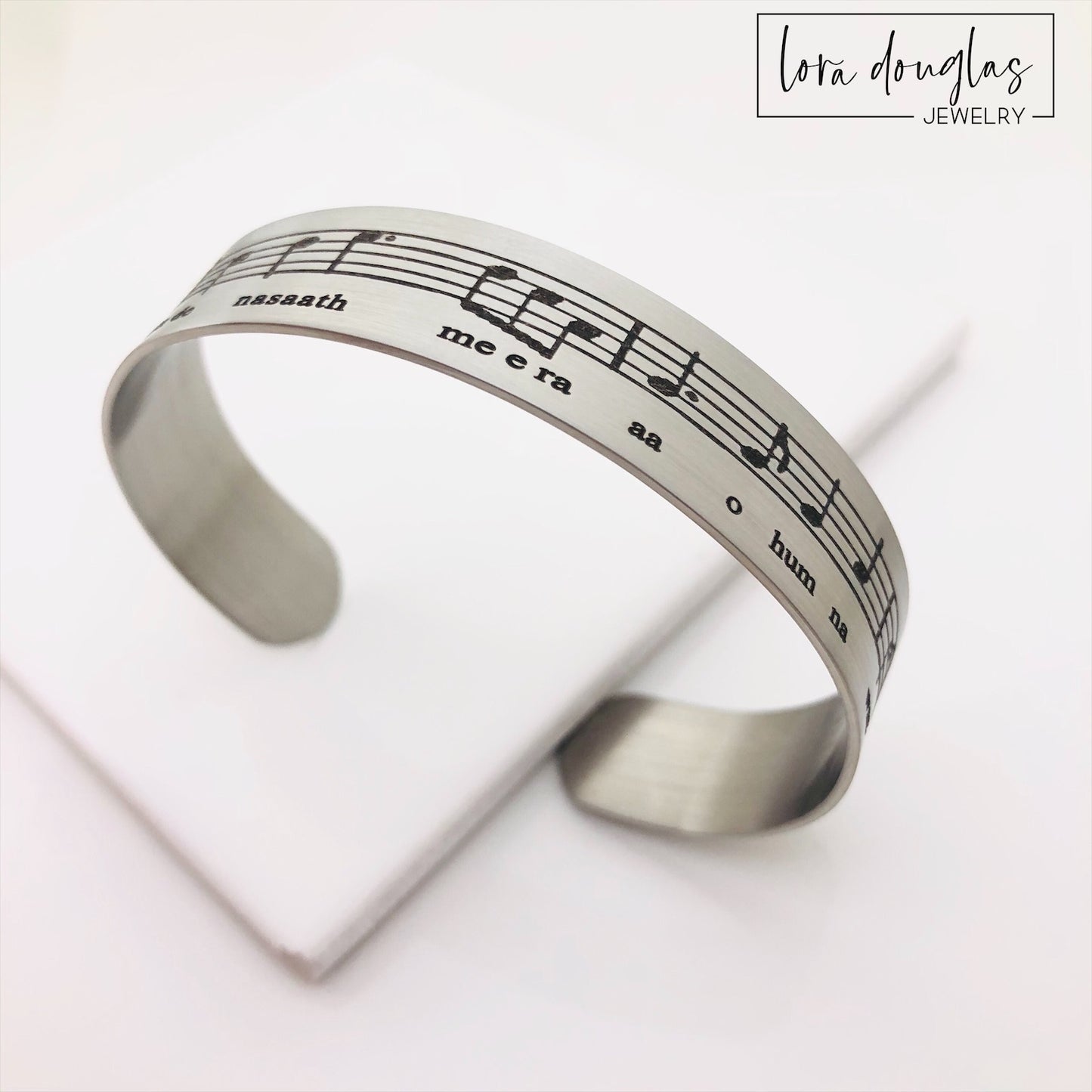 A stainless steel bracelet with sheet music engraved on it, displayed on a black surface with a green leafy element to the side.