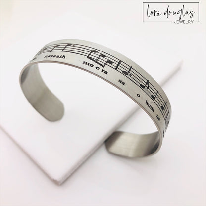 A stainless steel bracelet with sheet music engraved on it, displayed on a black surface with a green leafy element to the side.