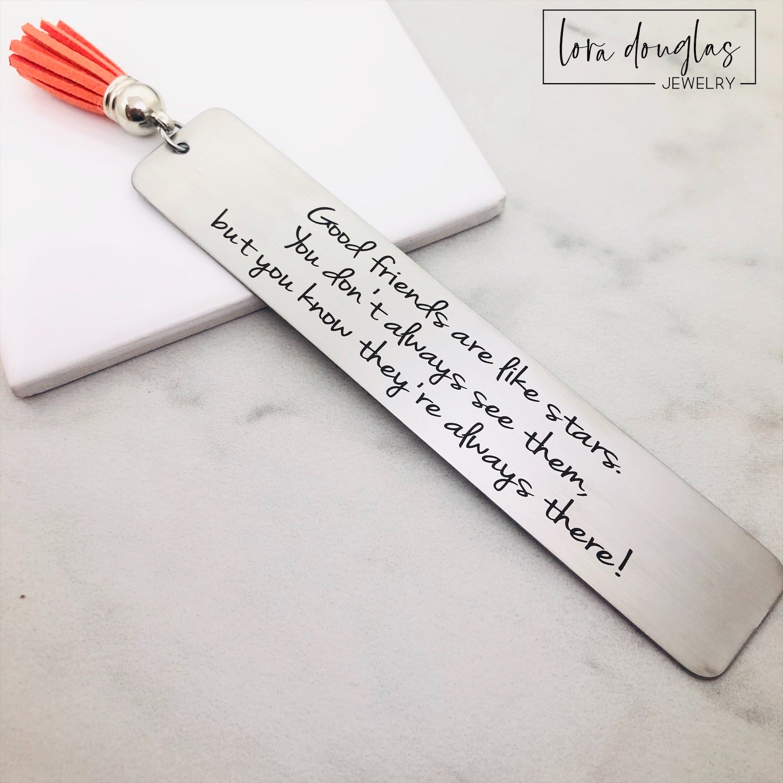 A stainless steel bookmark with an engraved message 'Good friends are like stars, you don't always see them but you know they're there.' placed on a book page, accompanied by a navy blue velvet drawstring pouch with a gift tag.