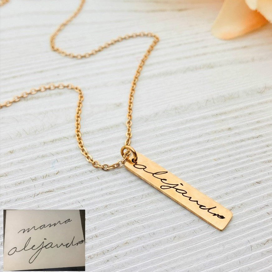 A gold stainless steel pendant with engraved handwriting, attached to an 18 inch chain with a lobster clasp, displayed against a white background with a blurred background hinting at a floral arrangement.