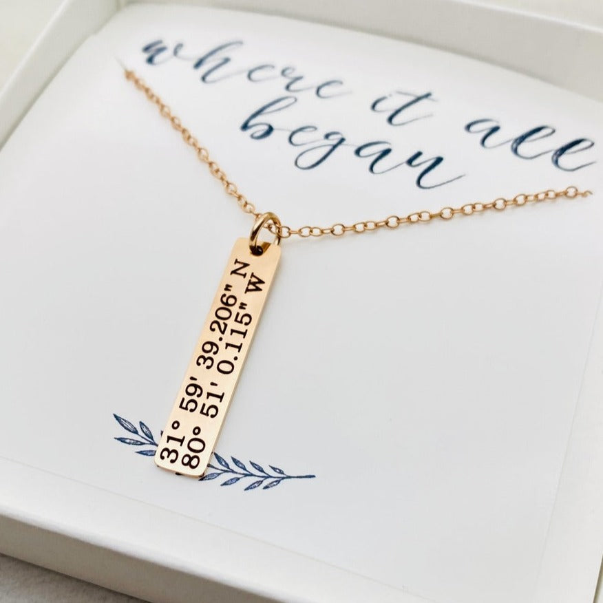 A rose gold necklace with a personalized engraving of a latitude and longitude on a pendant, displayed in a box with a blue and white design on the interior lining.