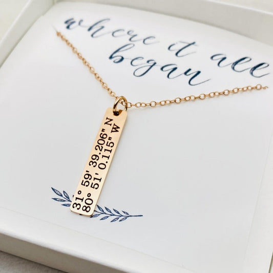 A rose gold necklace with a personalized engraving of a latitude and longitude on a pendant, displayed in a box with a blue and white design on the interior lining.