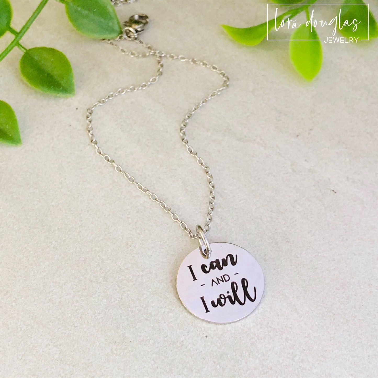 A silver-colored round pendant with the engraved text 'I CAN AND I WILL' on a chain, presented against a neutral background.
