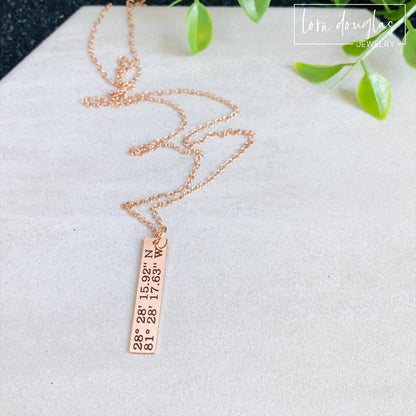 A rose gold necklace with a personalized engraving of a latitude and longitude on a pendant, displayed in a box with a blue and white design on the interior lining.