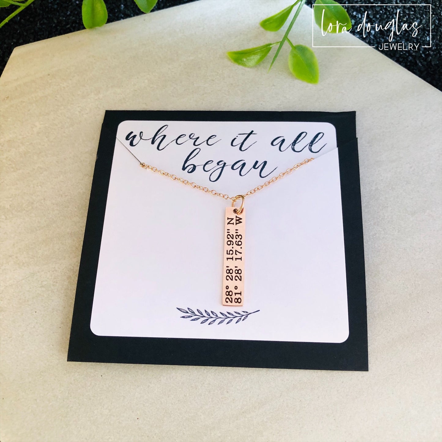 A rose gold necklace with a personalized engraving of a latitude and longitude on a pendant, displayed in a box with a blue and white design on the interior lining.