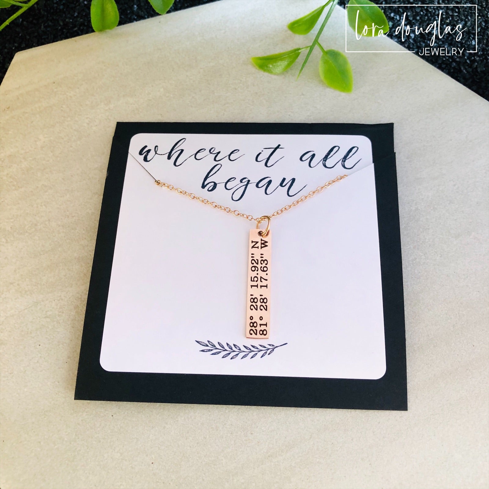 A rose gold necklace with a personalized engraving of a latitude and longitude on a pendant, displayed in a box with a blue and white design on the interior lining.