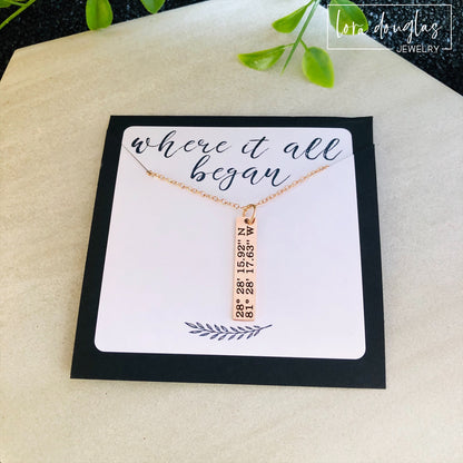 A rose gold necklace with a personalized engraving of a latitude and longitude on a pendant, displayed in a box with a blue and white design on the interior lining.