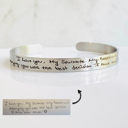 A stainless steel cuff bracelet with engraved handwriting on it, displayed on a light background.