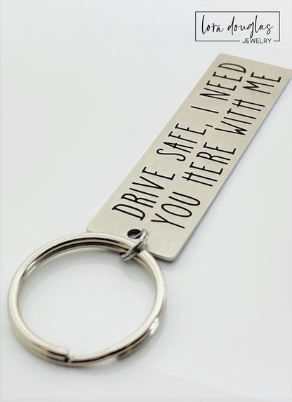 A metal key chain with the inscription 'DRIVE SAFE I NEED YOU HERE WITH ME' engraved on it.