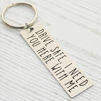 A metal key chain with the inscription 'DRIVE SAFE I NEED YOU HERE WITH ME' engraved on it.