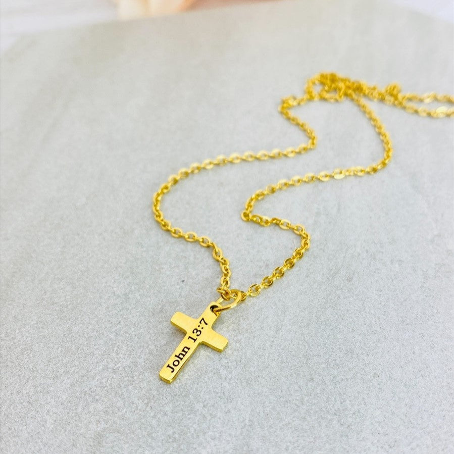 A gold stainless steel engraved cross necklace with a lobster clasp and a small cross charm, placed on a white surface