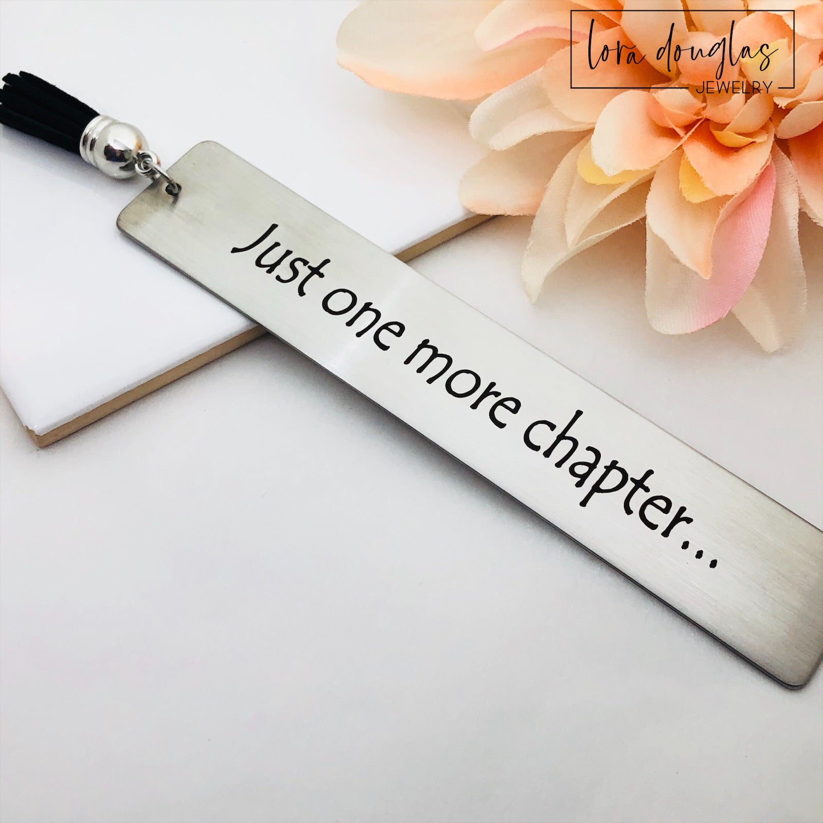 A stainless steel metal bookmark with the engraved text 'Just One More Chapter' and a tassel, placed on an open book.