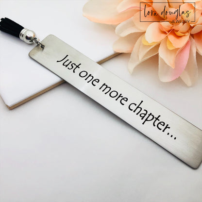 A stainless steel metal bookmark with the engraved text 'Just One More Chapter' and a tassel, placed on an open book.