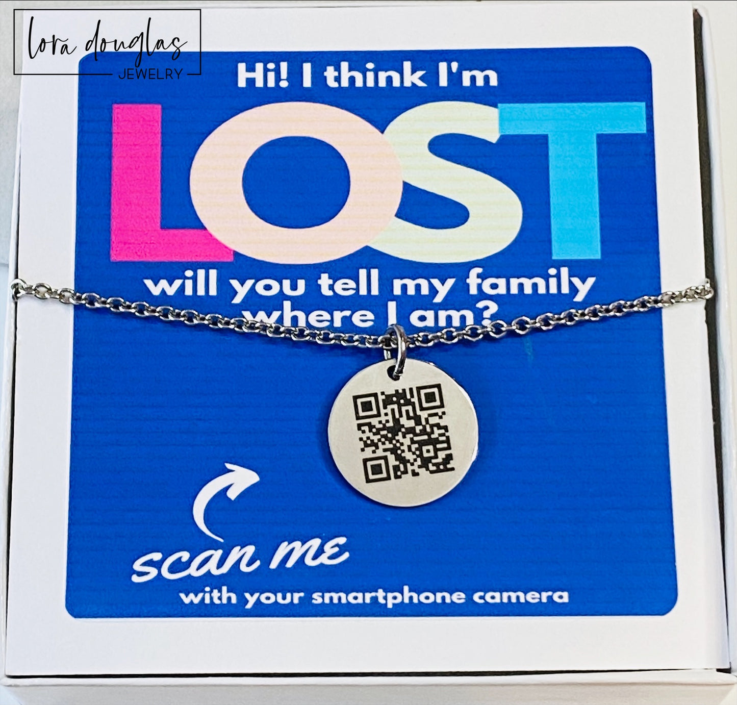 A QR code charm placed on a display stand, with a silver chain necklace that says 'Lost - will you tell my family I'm ok?' engraved on a blue disc.