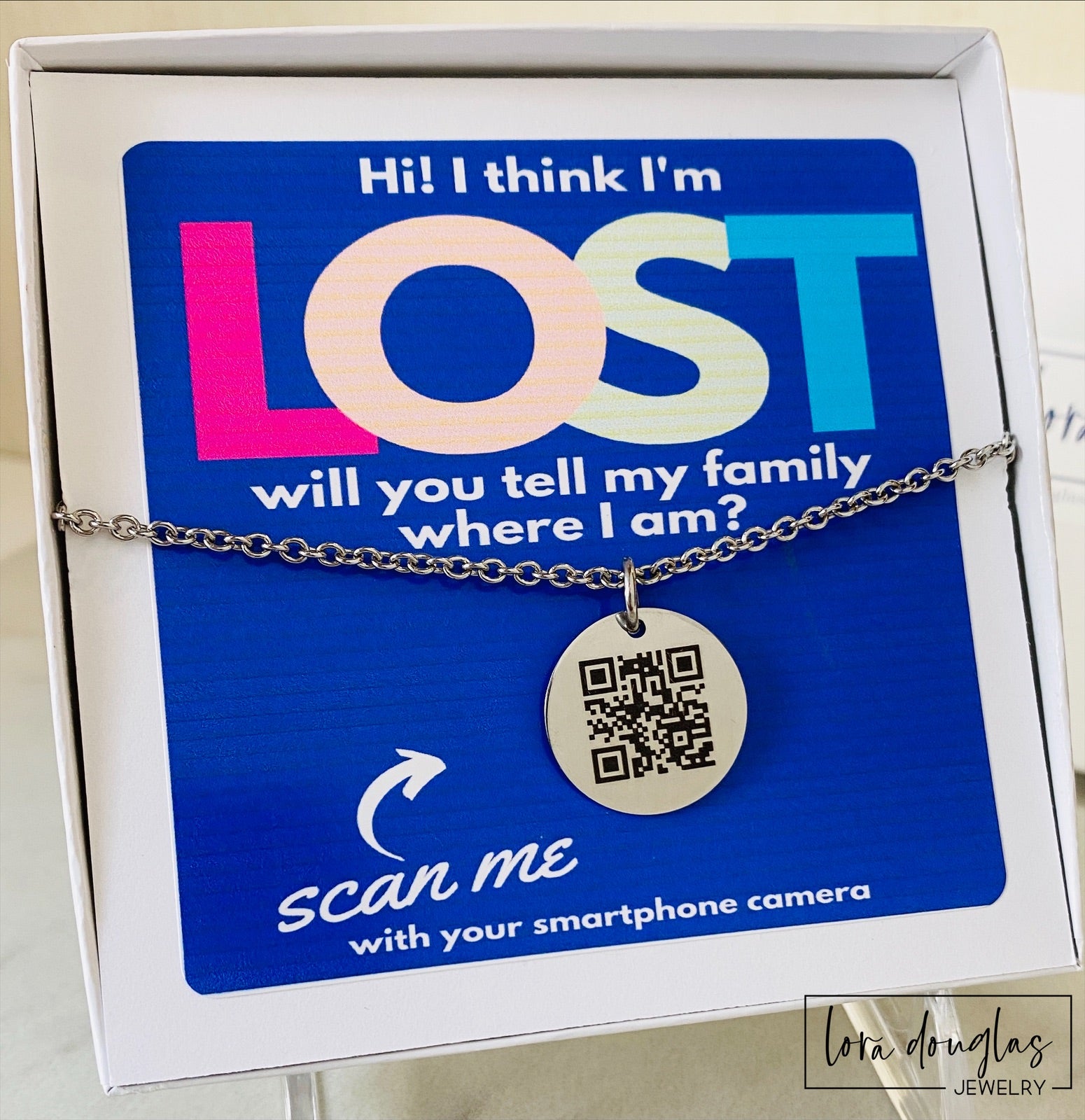 A QR code charm placed on a display stand, with a silver chain necklace that says 'Lost - will you tell my family I'm ok?' engraved on a blue disc.