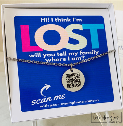 A QR code charm placed on a display stand, with a silver chain necklace that says 'Lost - will you tell my family I'm ok?' engraved on a blue disc.