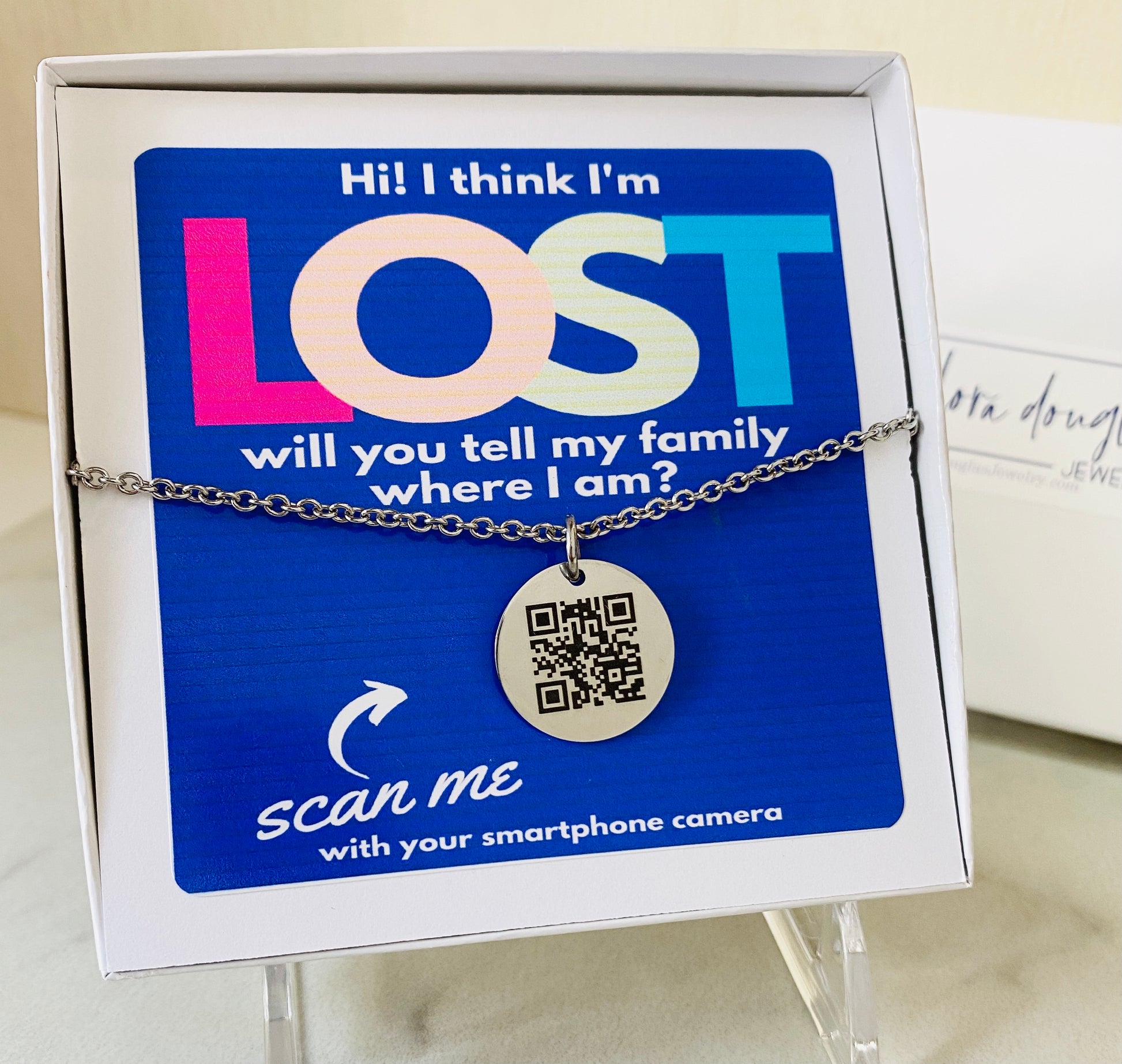 A QR code charm placed on a display stand, with a silver chain necklace that says 'Lost - will you tell my family I'm ok?' engraved on a blue disc.