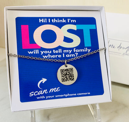A QR code charm placed on a display stand, with a silver chain necklace that says 'Lost - will you tell my family I'm ok?' engraved on a blue disc.