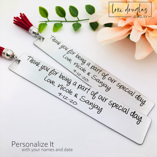 Two stainless steel bookmarks with personalized engraving, 'Thank you for being a part of our special day' and names, the other with a date. They are displayed on a white background with decorative tassels and flowers in the background.