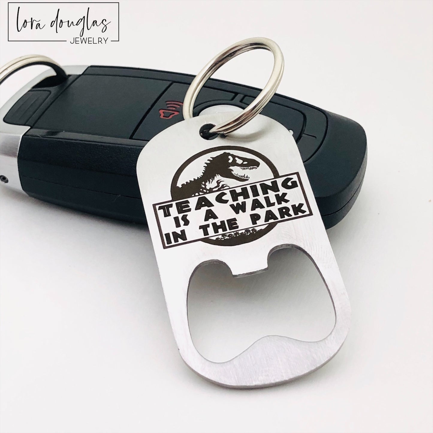 A stainless steel bottle opener with keychain featuring an engraving that says 'TEACHING IS A WALK IN THE PARK'.