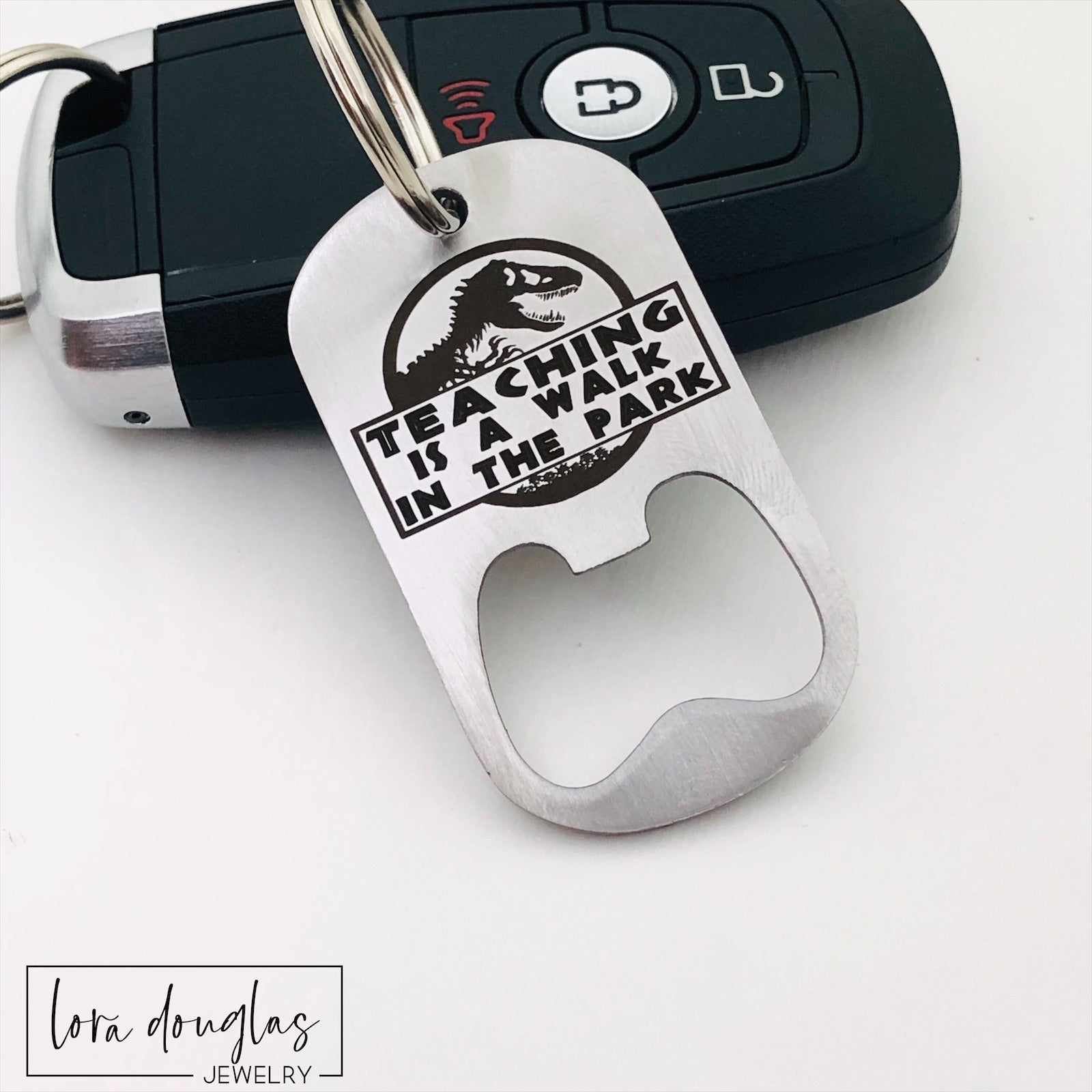 A stainless steel bottle opener with keychain featuring an engraving that says 'TEACHING IS A WALK IN THE PARK'.