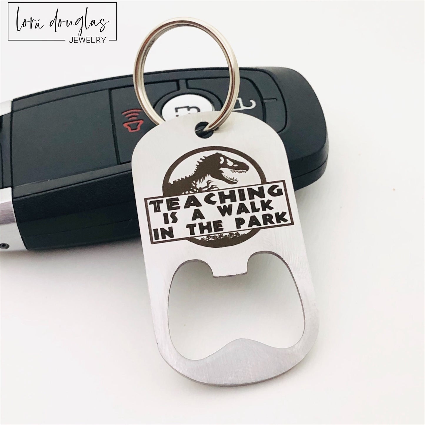 A stainless steel bottle opener with keychain featuring an engraving that says 'TEACHING IS A WALK IN THE PARK'.