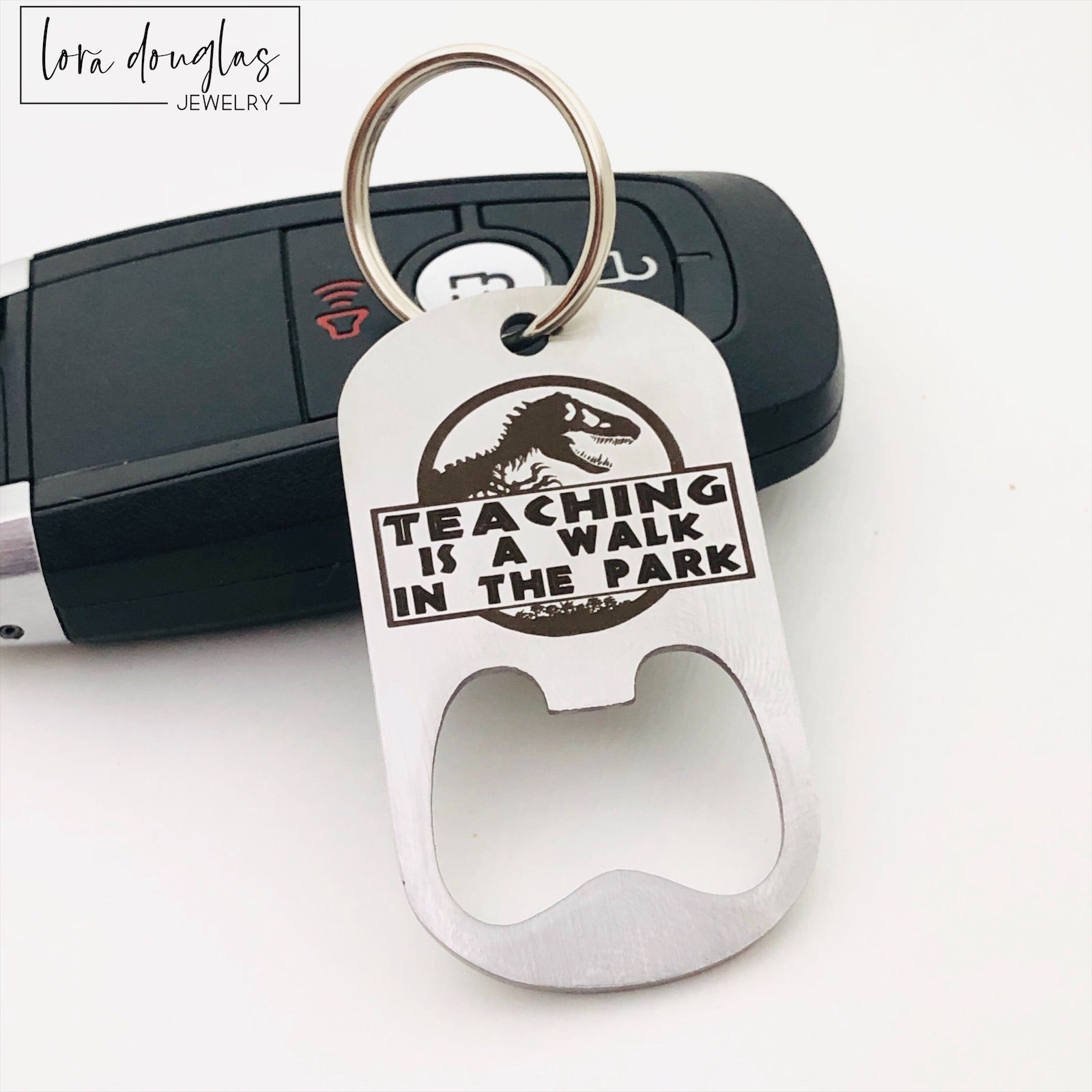 A stainless steel bottle opener with keychain featuring an engraving that says 'TEACHING IS A WALK IN THE PARK'.