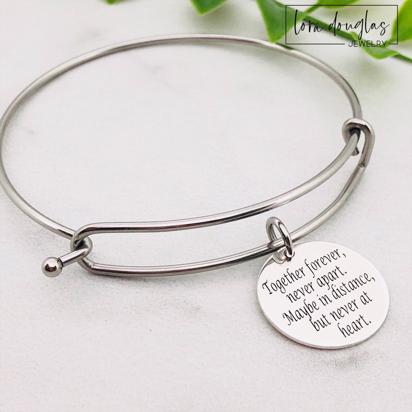 A stainless steel adjustable bangle bracelet with a round pendant that has engraved text 'Together Forever, Never Apart, Maybe in Distance, But Never at Heart'.