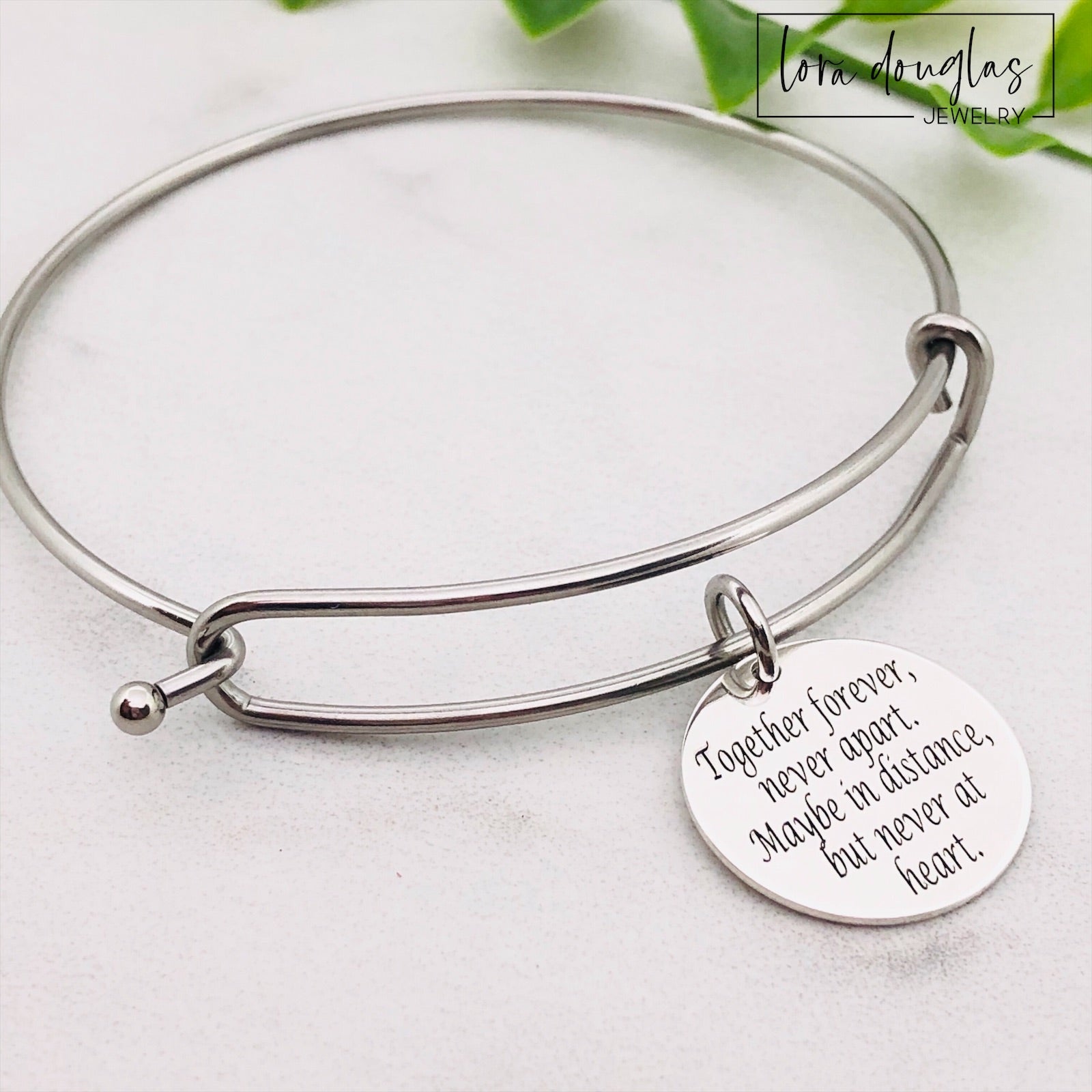 A stainless steel adjustable bangle bracelet with a round pendant that has engraved text 'Together Forever, Never Apart, Maybe in Distance, But Never at Heart'.