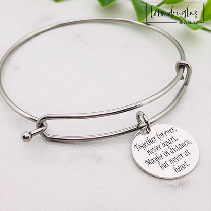 A stainless steel adjustable bangle bracelet with a round pendant that has engraved text 'Together Forever, Never Apart, Maybe in Distance, But Never at Heart'.