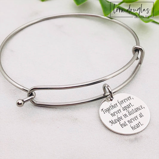 A stainless steel adjustable bangle bracelet with a round pendant that has engraved text 'Together Forever, Never Apart, Maybe in Distance, But Never at Heart'.