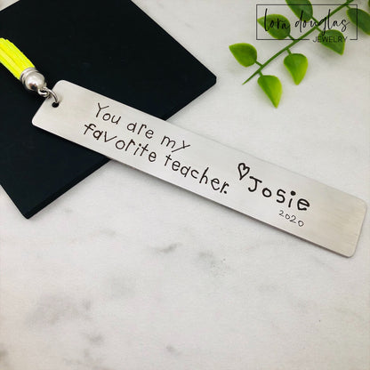 Two stainless steel bookmarks with engraved handwriting, with tassels, displayed on a wooden surface.