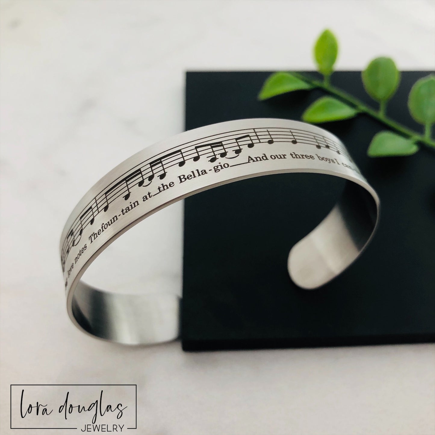 A stainless steel bracelet with sheet music engraved on it, displayed on a black surface with a green leafy element to the side.