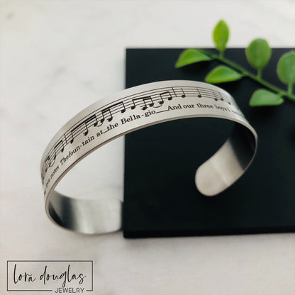 A stainless steel bracelet with sheet music engraved on it, displayed on a black surface with a green leafy element to the side.