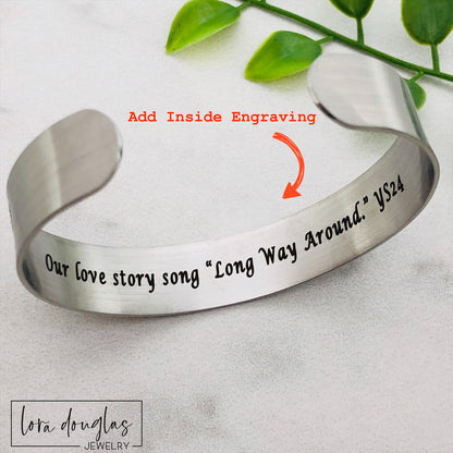 A stainless steel bracelet with sheet music engraved on it, displayed on a black surface with a green leafy element to the side.