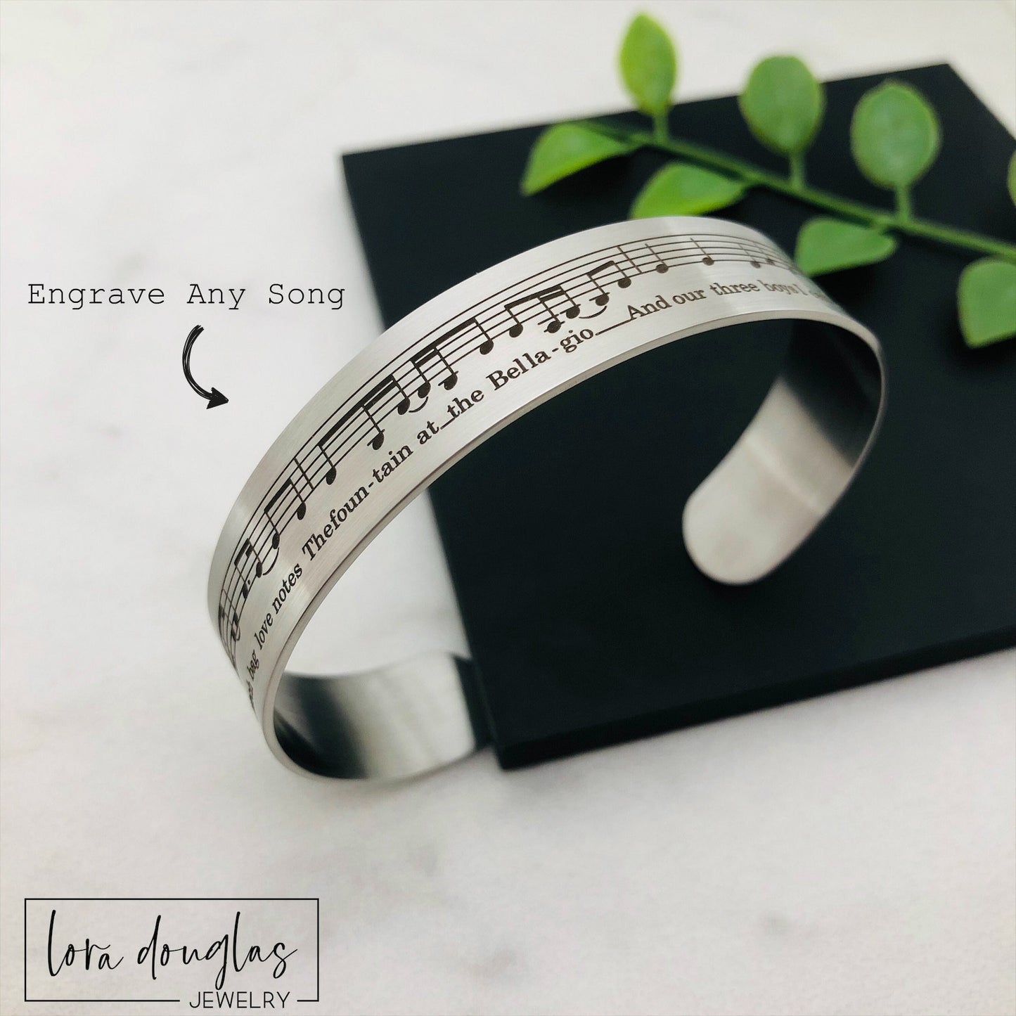 A stainless steel bracelet with sheet music engraved on it, displayed on a black surface with a green leafy element to the side.