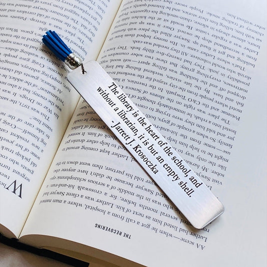 A stainless steel tassel bookmark with an engraved quote 'The library is the heart of the school...' placed on an open book.