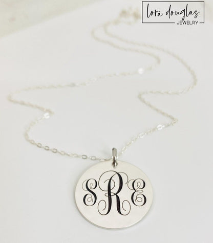A silver monogram necklace with an engraved initial pendant laid out on a marble surface.