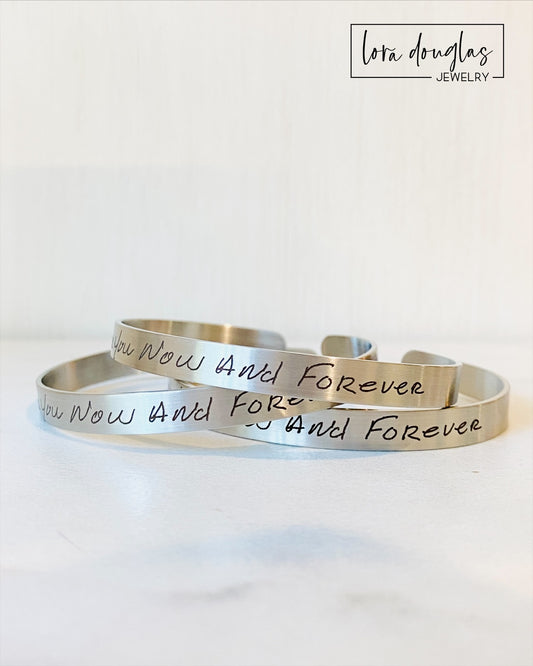 A stainless steel cuff bracelet with engraved handwriting on it, displayed on a light background.