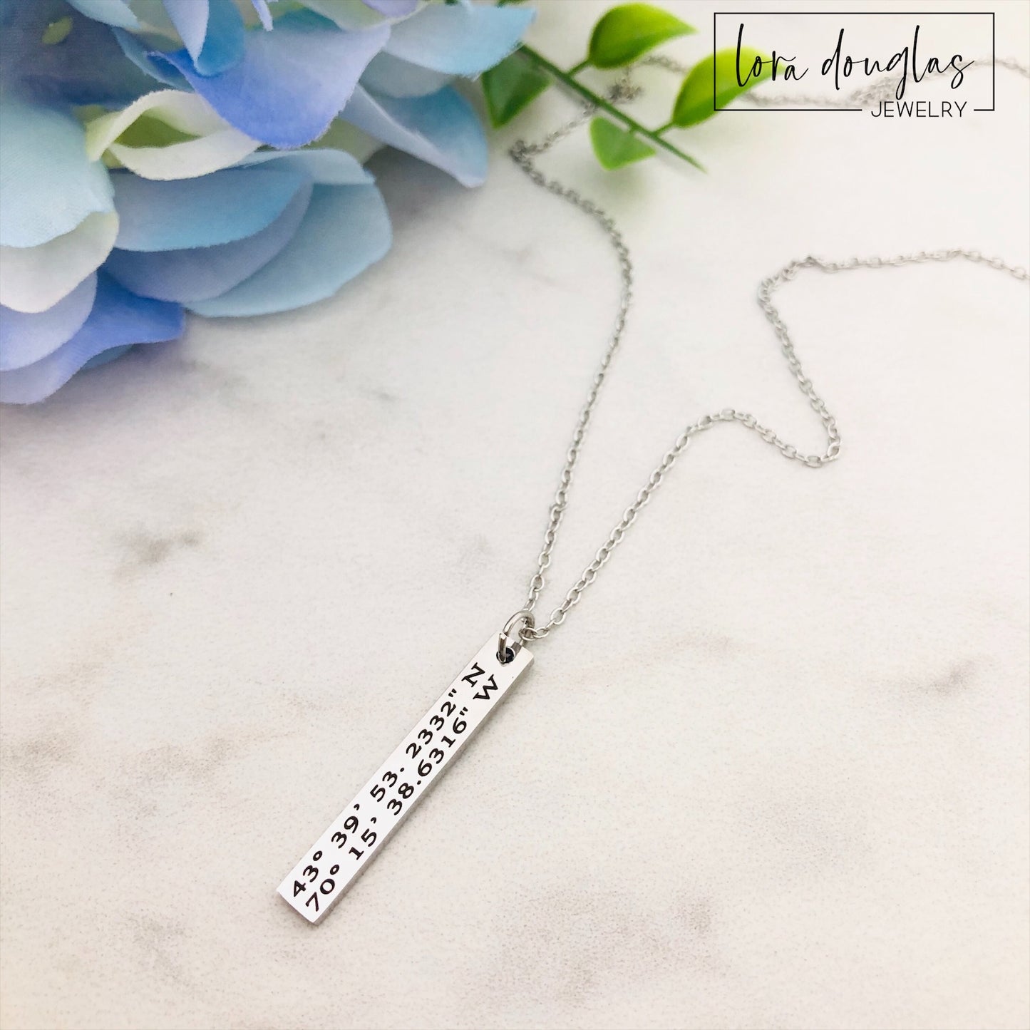 A custom coordinates necklace with a silver stainless steel pendant and chain, displayed against a floral background.