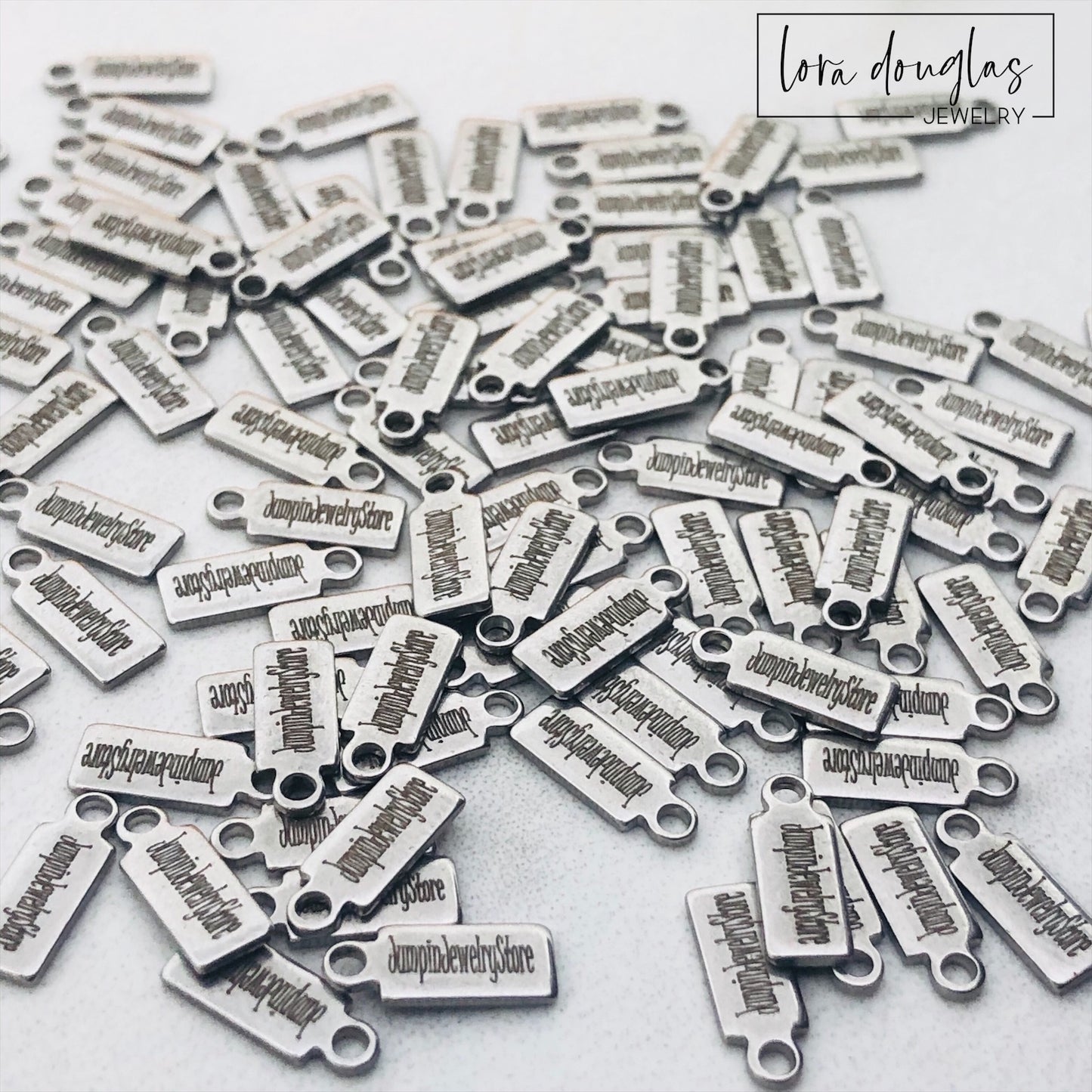A collection of stainless steel jewelry tags with laser engraved logos and text on a white surface.