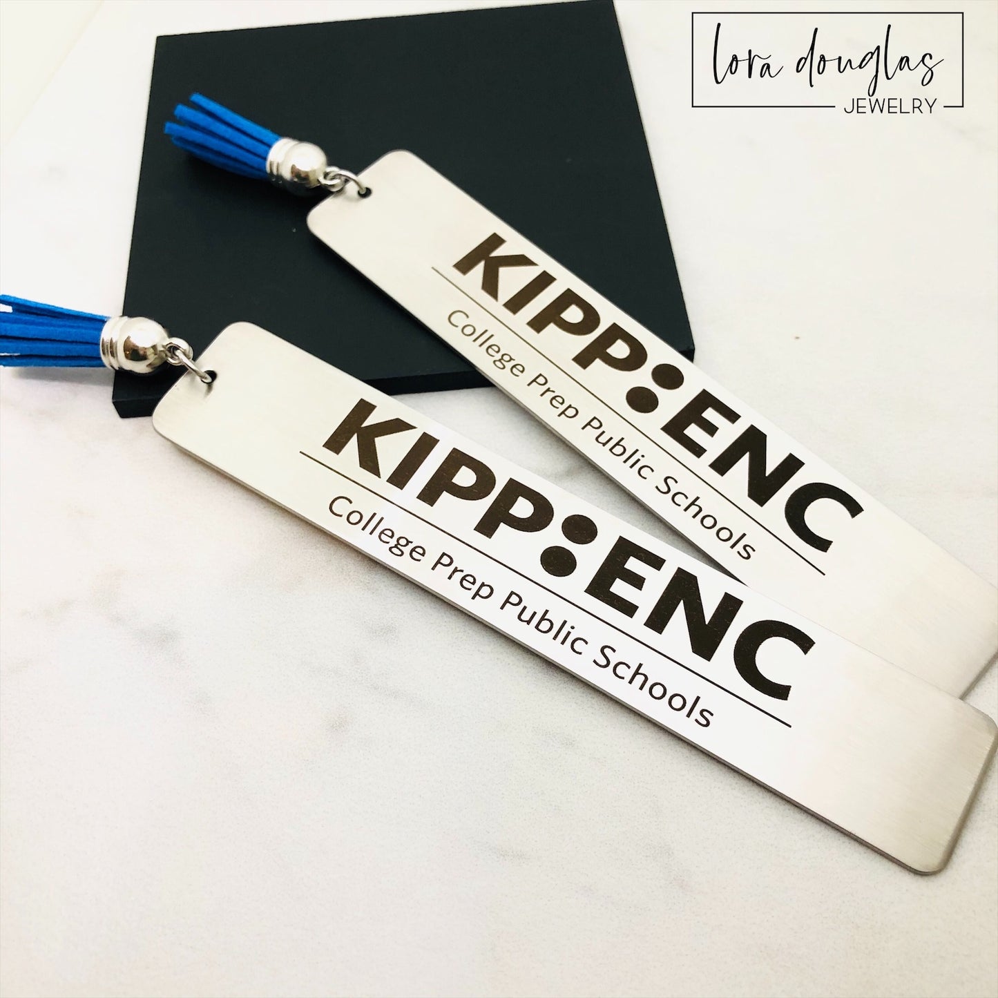 Two stainless steel bookmarks with custom engravings, one displaying a logo and the other a graphic. Both have blue tassels and are presented on a white surface.