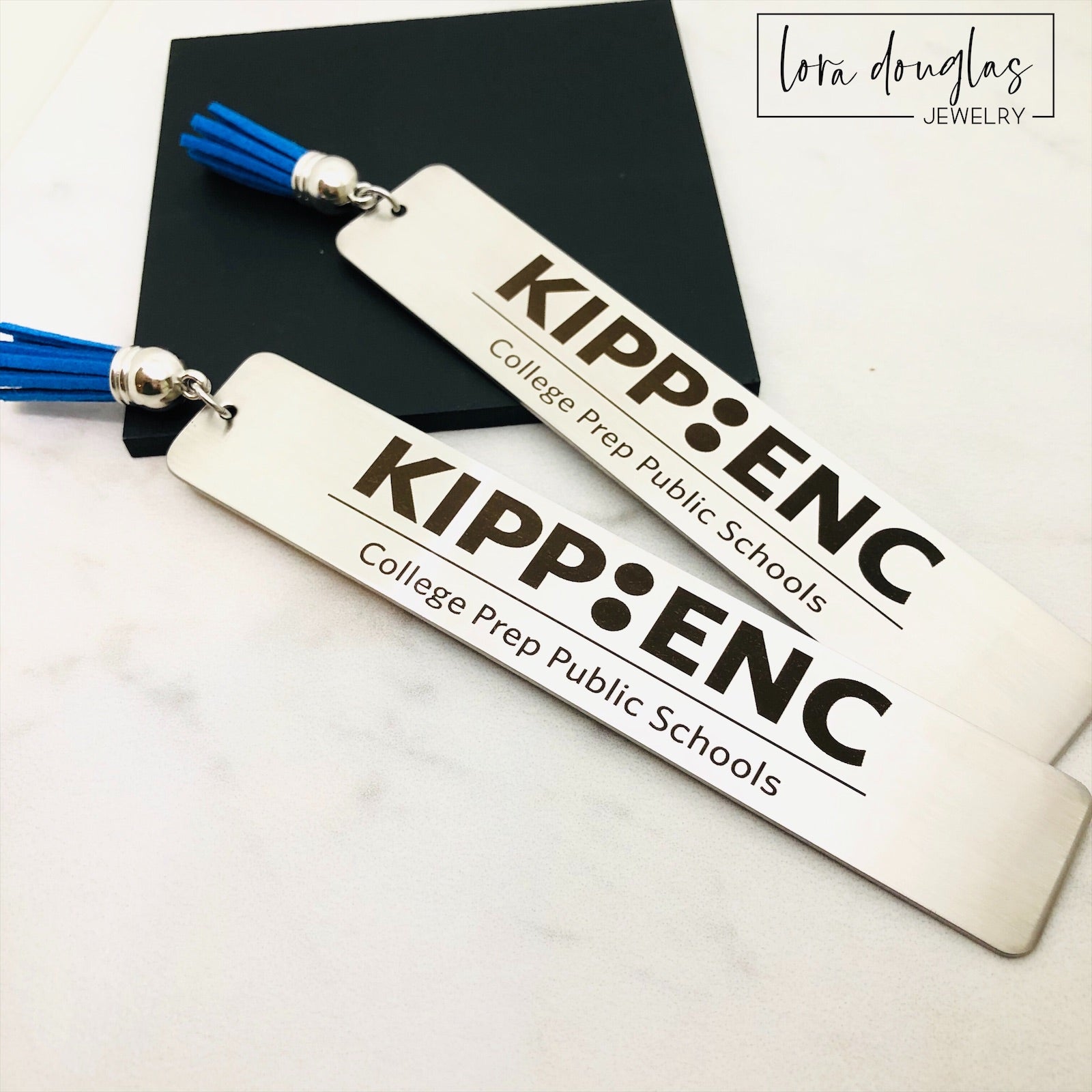 Two stainless steel bookmarks with custom engravings, one displaying a logo and the other a graphic. Both have blue tassels and are presented on a white surface.