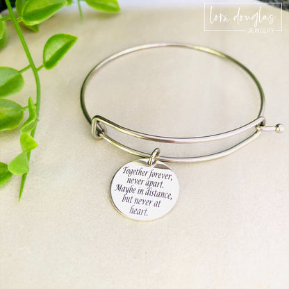 A stainless steel adjustable bangle bracelet with a round pendant that has engraved text 'Together Forever, Never Apart, Maybe in Distance, But Never at Heart'.
