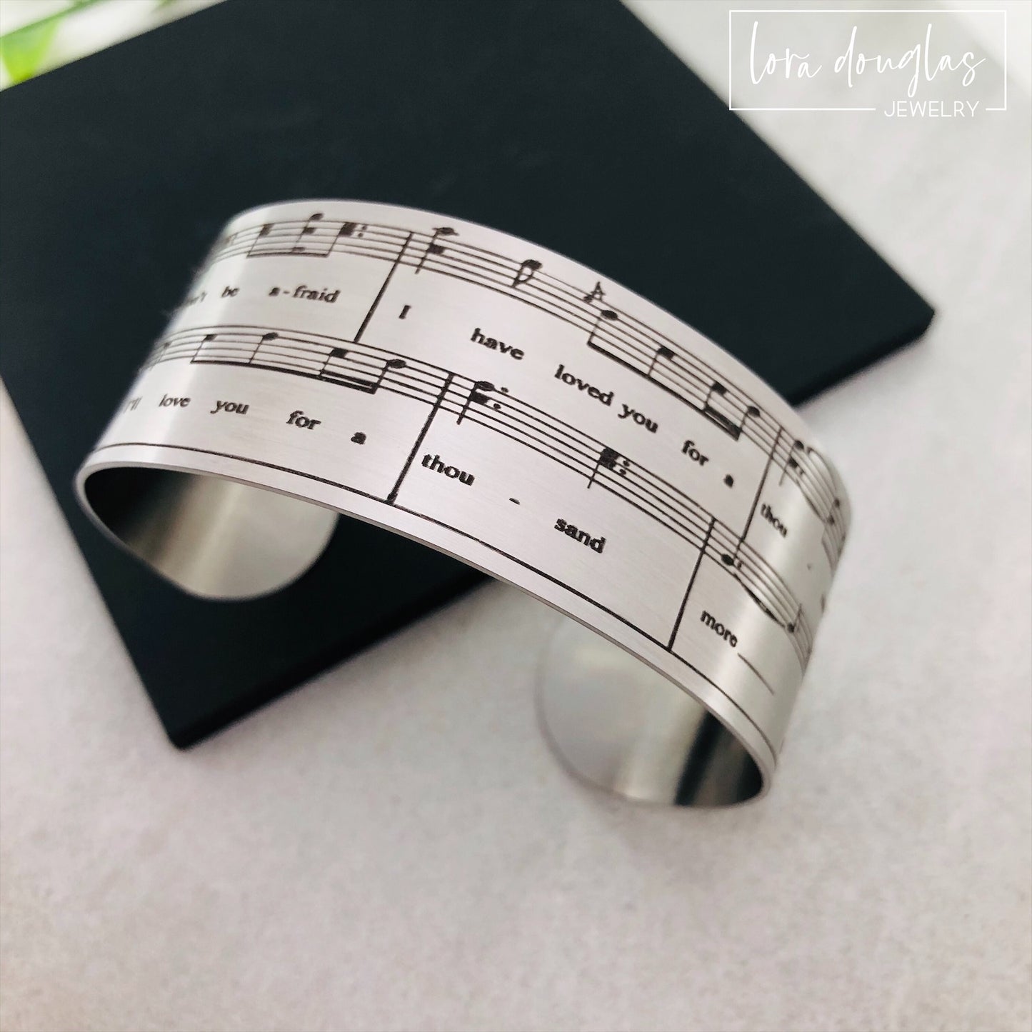 A stainless steel bracelet with sheet music engraving, displayed on a background of sheet music.